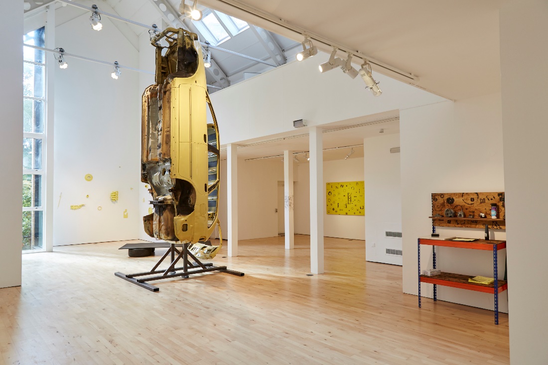 Mustard-coloured car frame on a stand in an art gallery space.