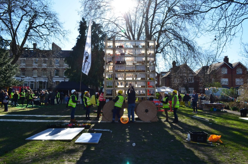 A group of people wearing hi-viz jackets and hard hats surround a tall wooden grid-like structure on wheels with lots of milk bottles hanging from it in a suburban park.