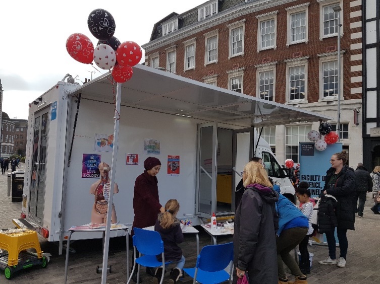 A small group of people are doing an interactive activity in front of a white van with a canopy in Kingston's Marketplace.