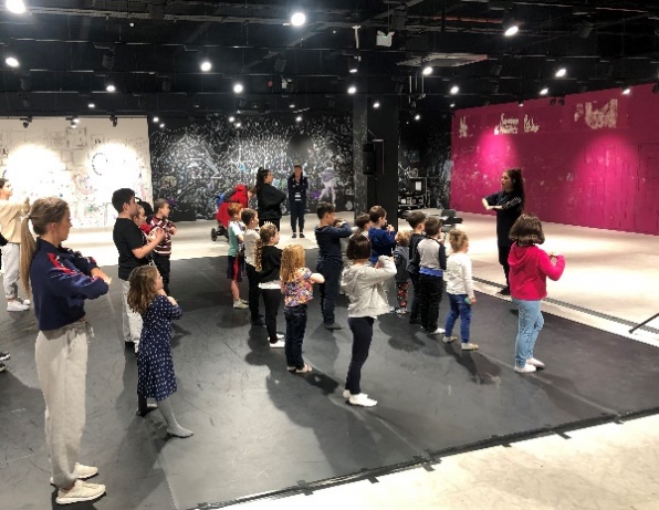 Group of approximately 20 children and young people in rows facing an instructor teaching dance moves or actions in an empty retail space.