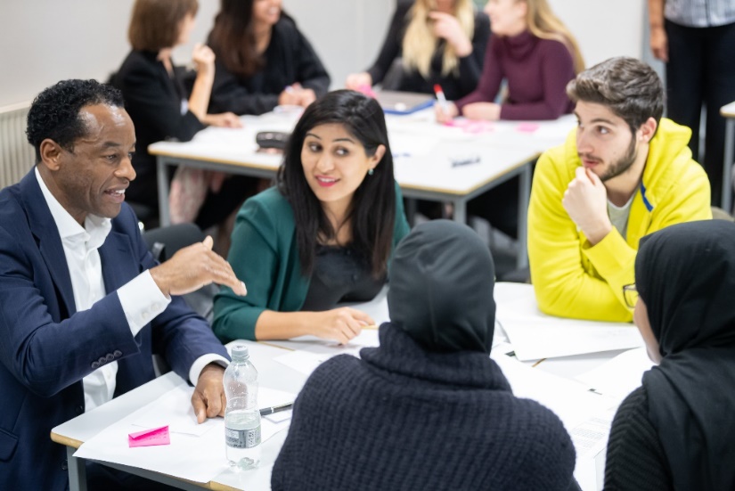Two tables with four to five people sitting round discussing a topic. Table in foreground includes an enterprise mentor giving feedback.