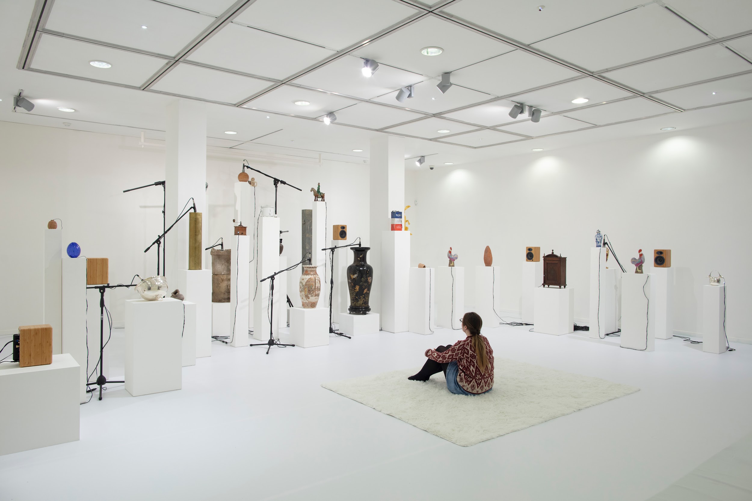 An image of an art gallery space, the walls, floor and ceiling are white. A range of objects sit on different sized plinths, microphones mounted on stands are positioned next to the objects. A woman sits on the floor on a white rug in front of the display of objects.