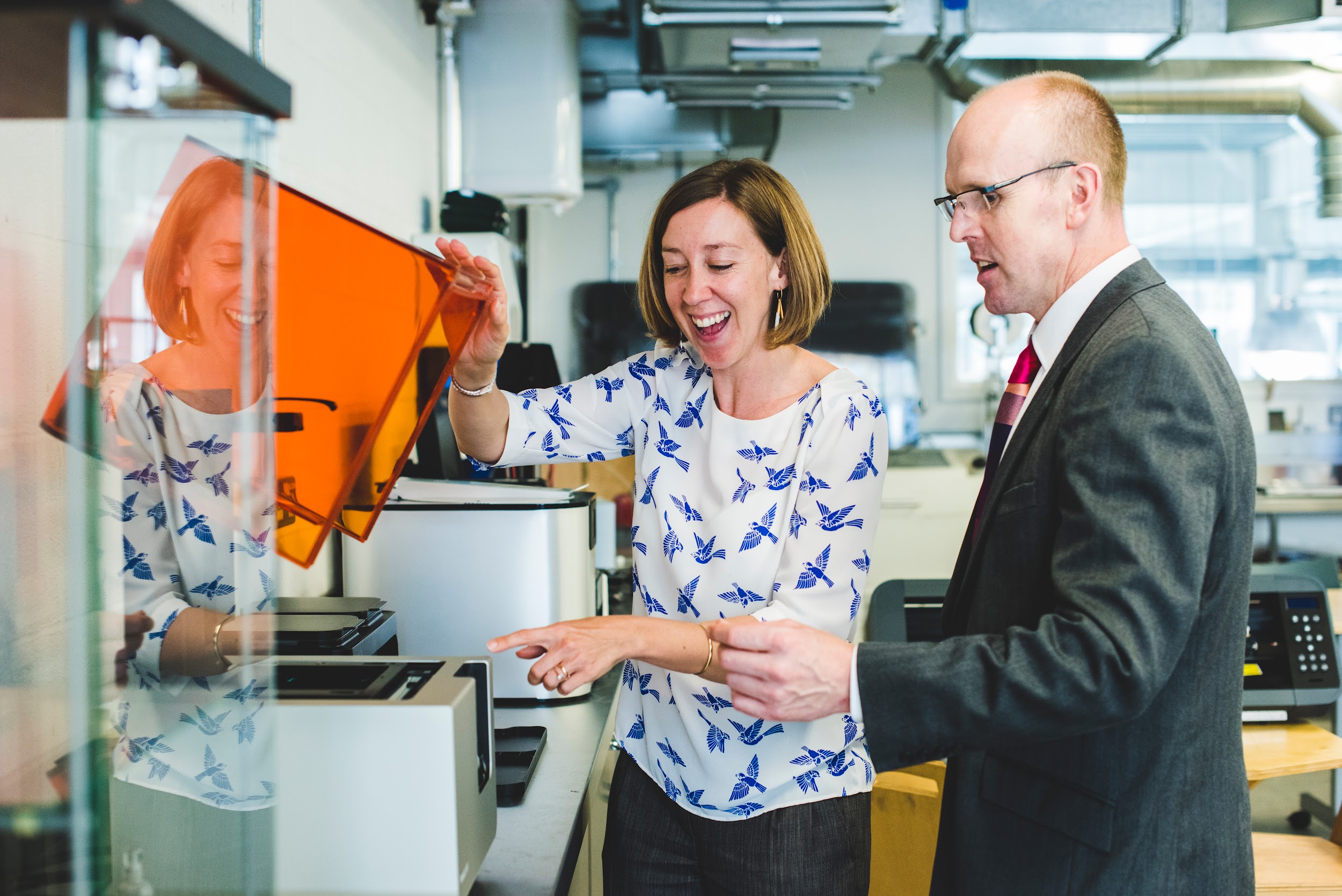 A man and a woman stand in front of a fabrication machine. The woman is smiling and holds the orange plastic lid of the machine while pointing.