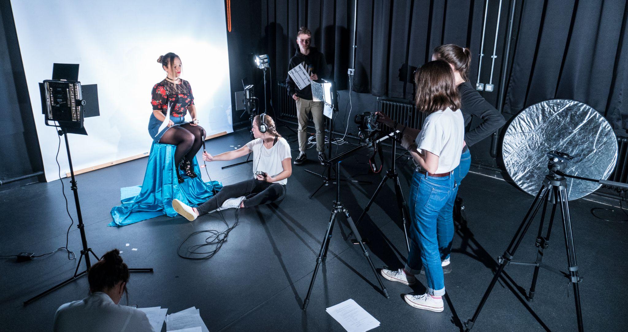 An image of a photography studio. A model sits on a chair draped in blue fabric, a persons wearing headphones sits on the floor next to the woman, another person stands in the background holding a clapper board. Two more women stand behind a camera which is pointed at the model.