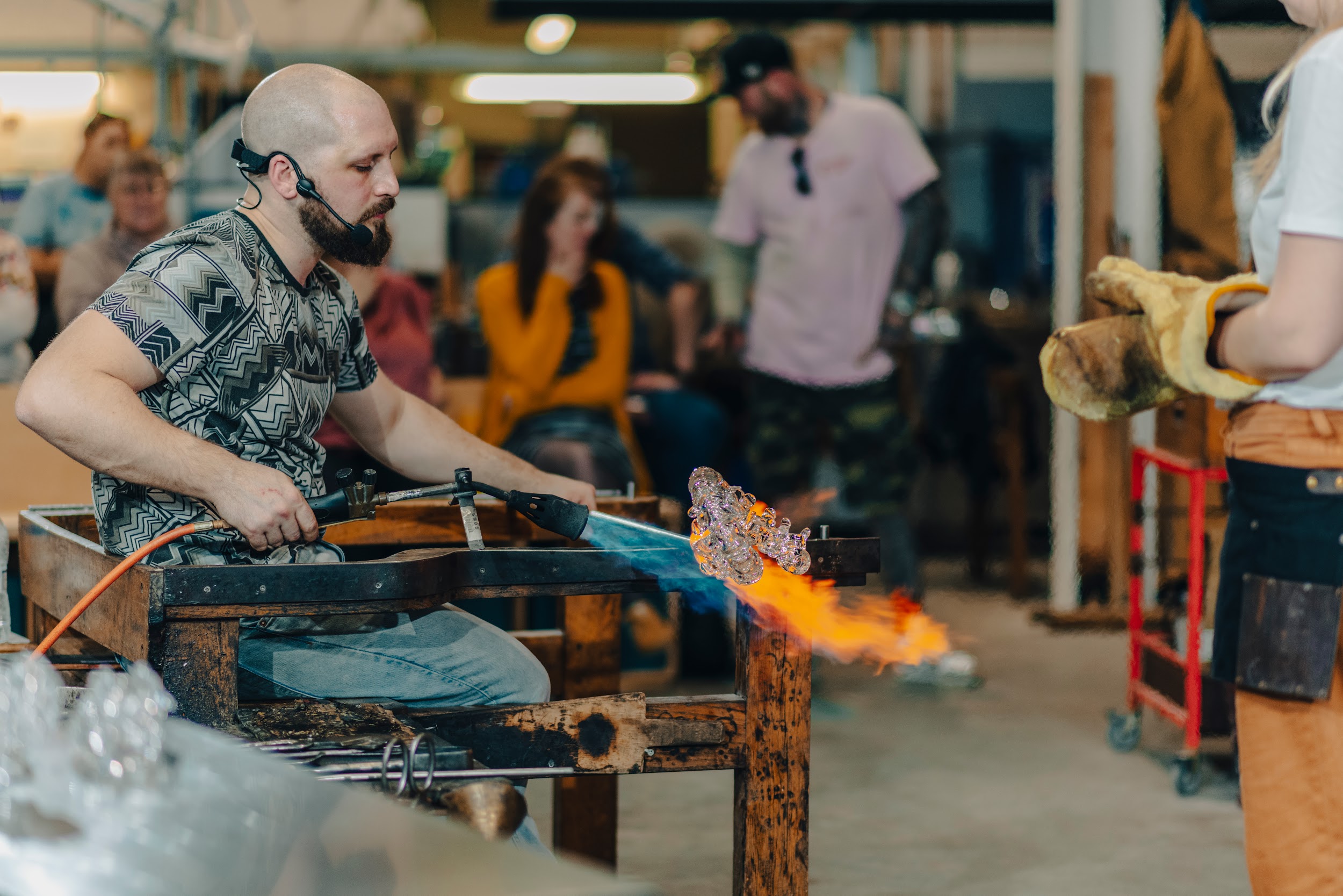 An image of a man sitting at a work bench, the man is holding a blow torch directed at a piece of hot glass. There are onlookers in the background.