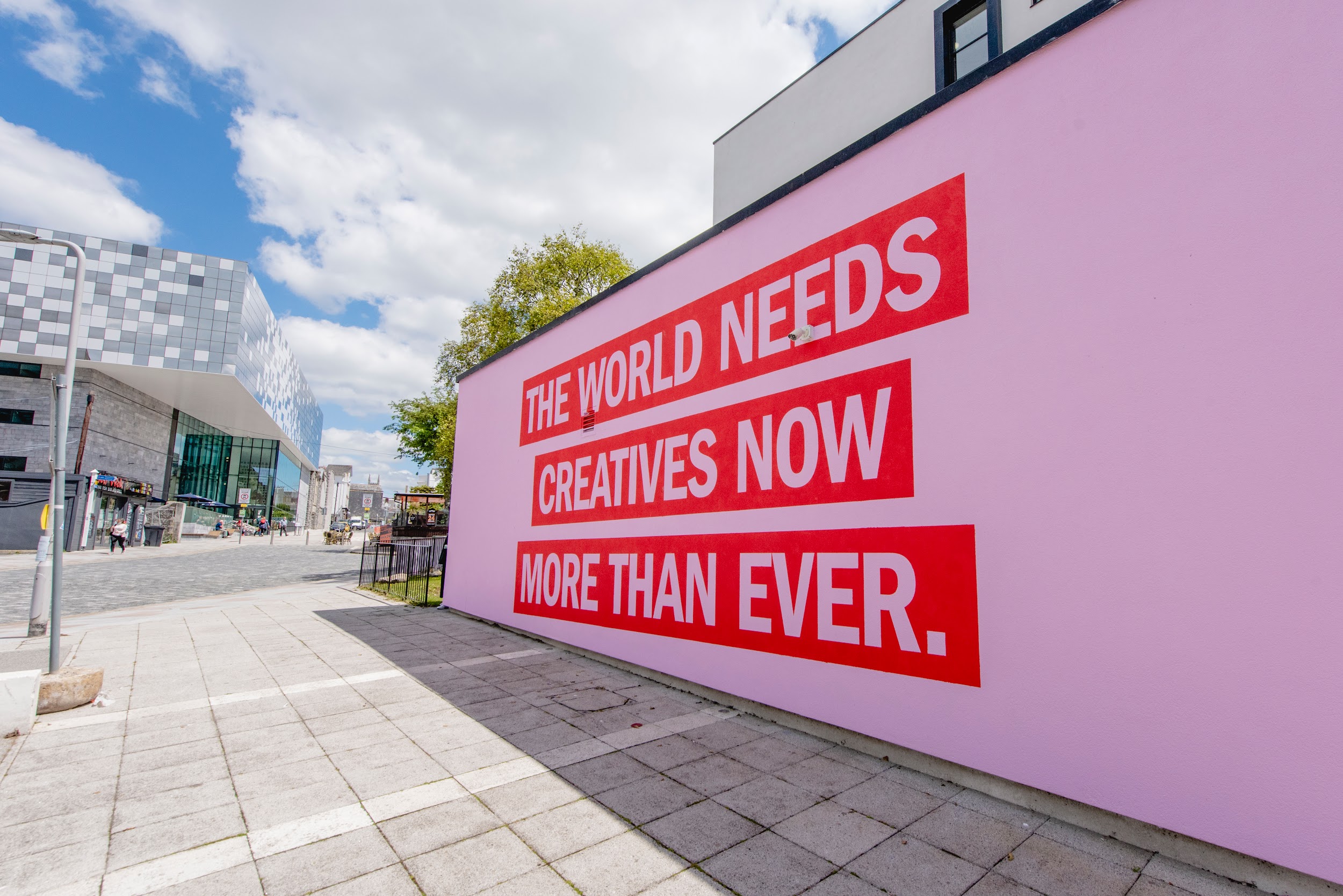 A close image of the exterior of Arts University Plymouth focusing on the words 'The world needs creatives now more than ever' in dark pink capital letters against a light pink background. In the back of the image you can see The Box Plymouth.