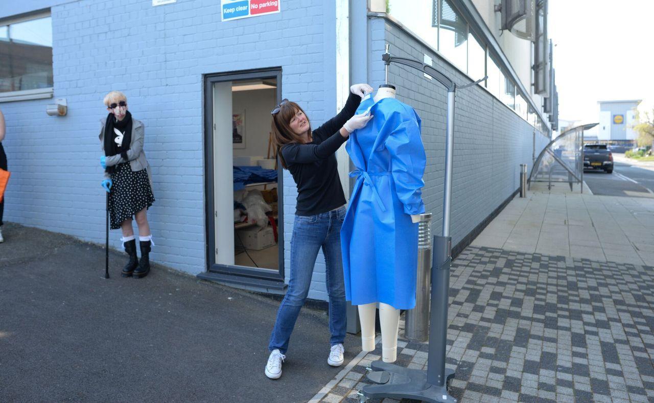 An image of a woman standing outside a building next to a dressmakers mannequin. The mannequin is wearing a piece of blue personal protective equipment. The woman us altering the blue piece of clothing on the dressmakers mannequin. Another woman looks on.