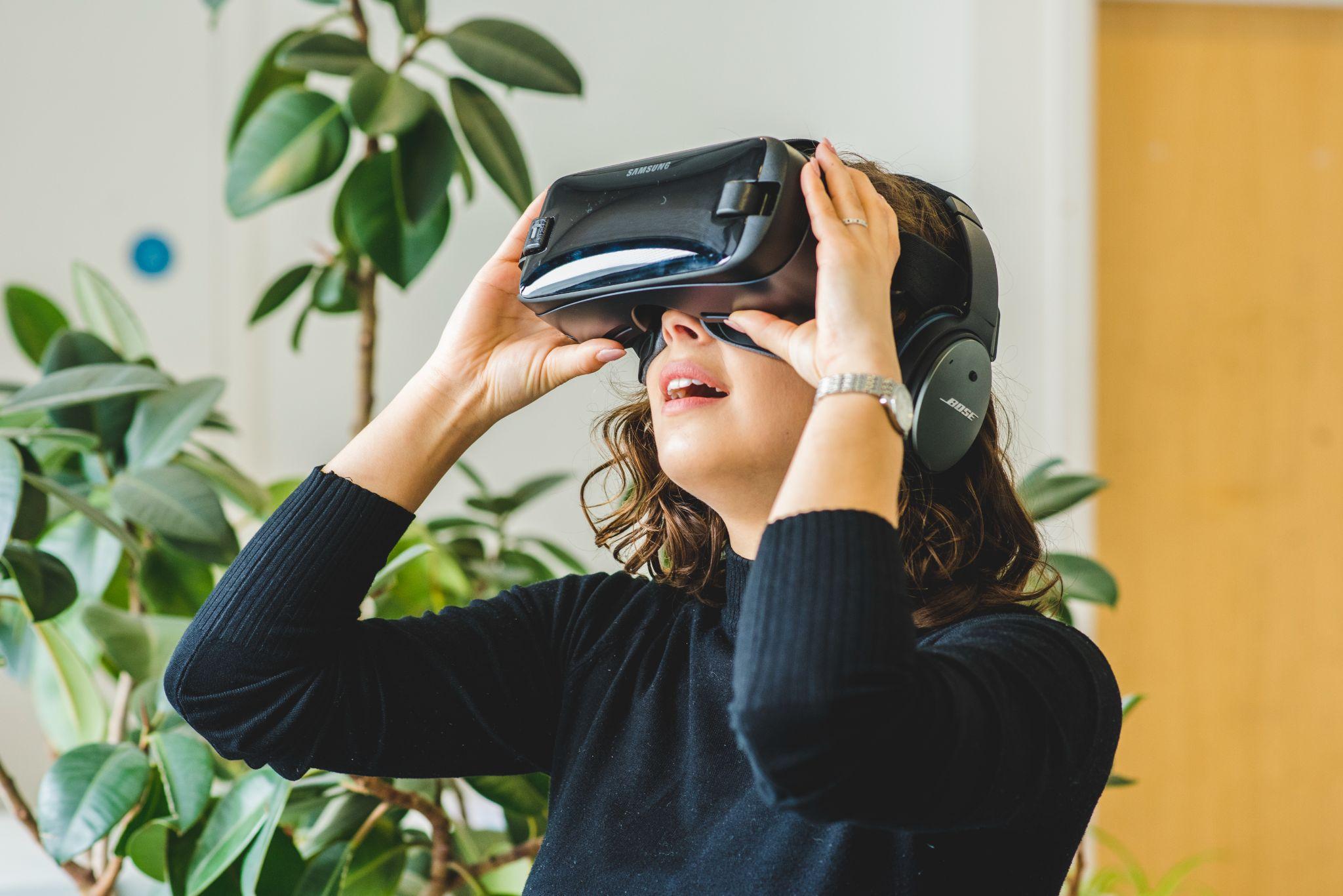 An image of a woman wearing a pair of virtual reality goggles. The woman has her hands on the goggles as she looks upwards.