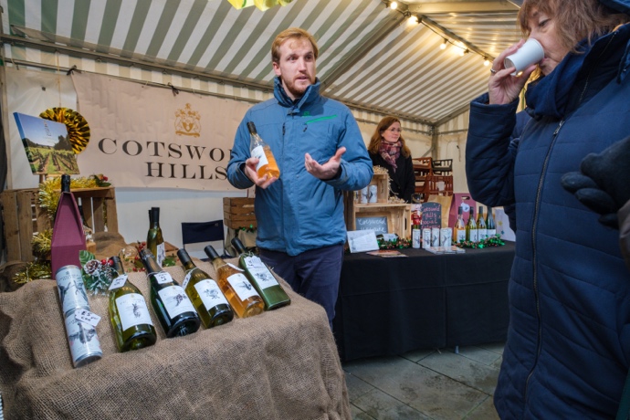Student chatting to a customer whilst holding a bottle of Cotswold Hills wine at Gloucester Christmas Market