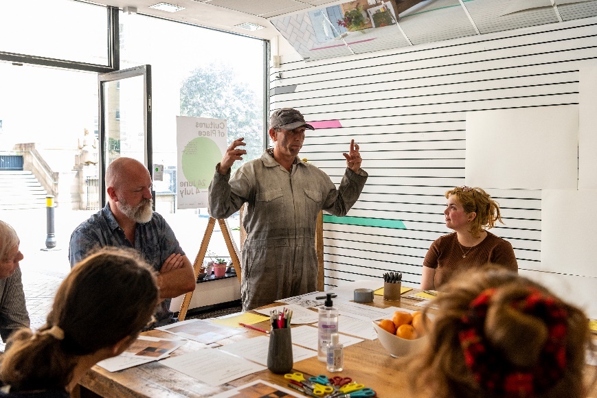 Photograph showing a presenter talking to a table of people. Presenter wearing a dirty boilersuit