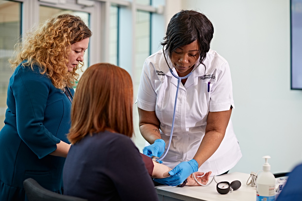 Standing figure dressed as a nurse taking the pulse of a seated person, back of head to camera. A third person, standing is watching on the left