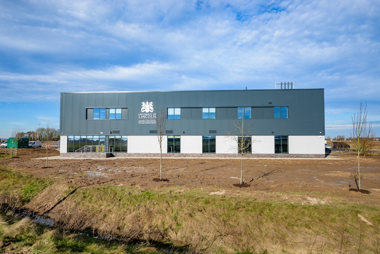 A photograph of the Centre of Excellence in Agri-Food Technologies building against a blue sky with mottled clouds, with newly planted young trees in the foreground.