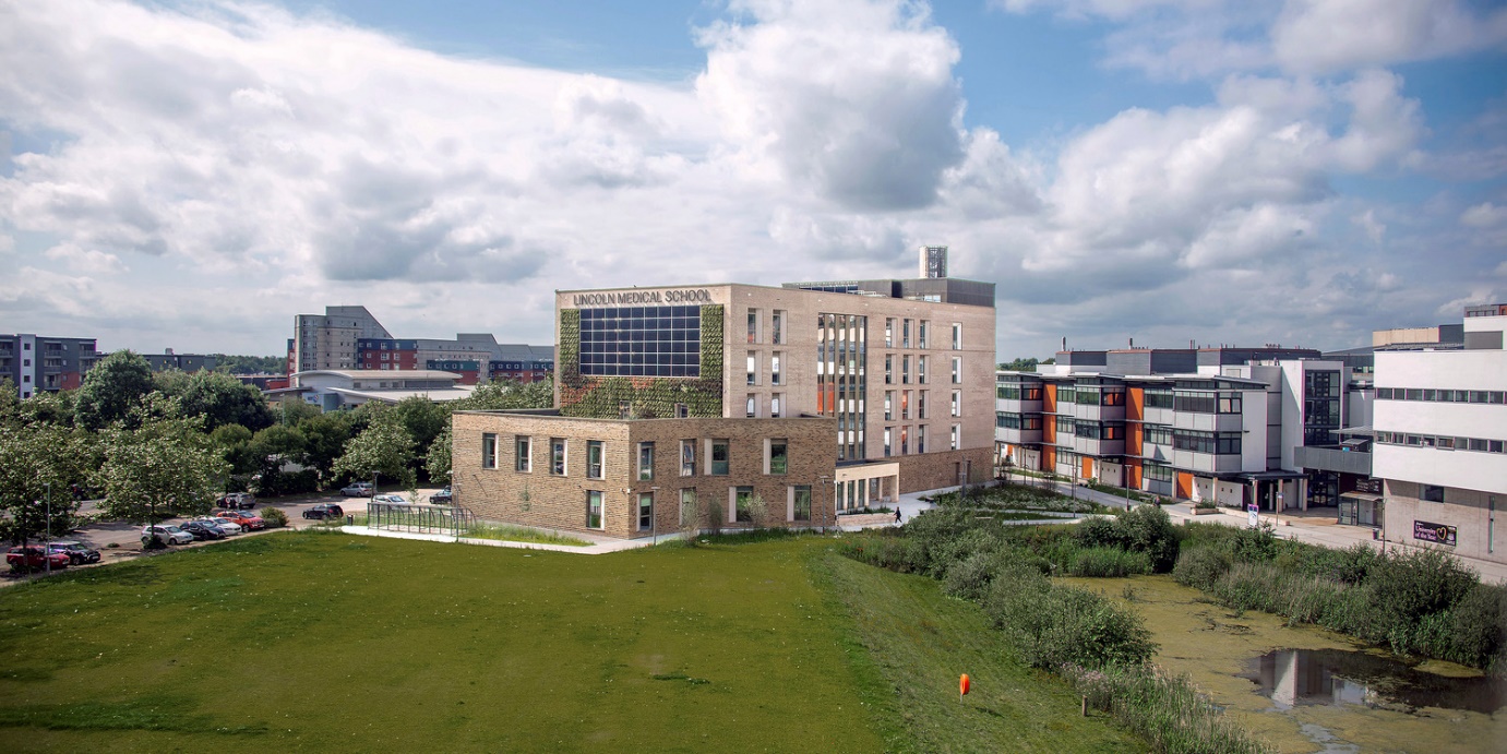 A picture containing grass, sky, outdoor, green and at the centre the Lincoln Medical School building with a living wall covered in plants and solar panels. Description automatically generated