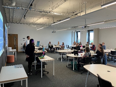 A group of people in a room around tables, listening to a speaker