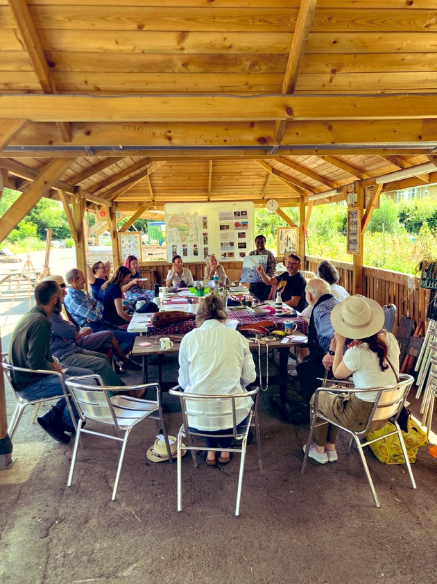 A group of about 20 people from the local community sit under cover, outside, around a table, talking