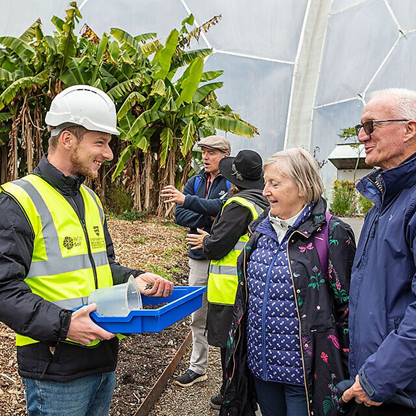 A researcher, with his-vis and a hard hat, holding a blue tray filled with soil talking with a man and woman.