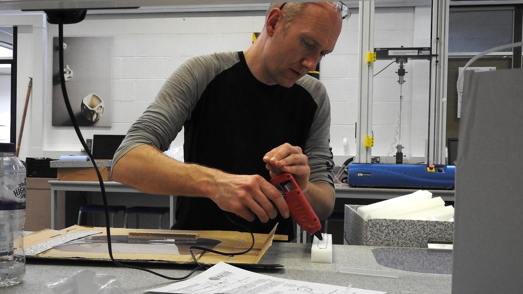 A white man, wearing a dark top, is holding a hot glue gun onto a piece of white material. In front of the man is a table with bits of wood, plastic and paper on it.