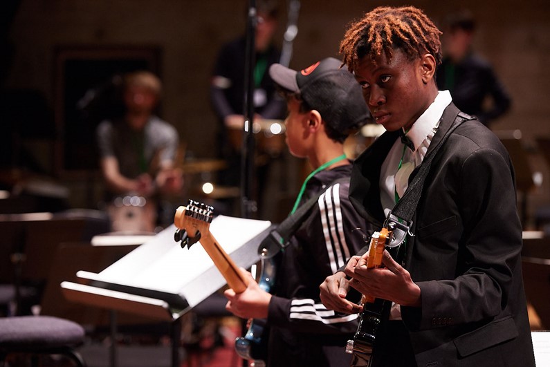 Picture of two boys playing electric guitars with drummers in the background