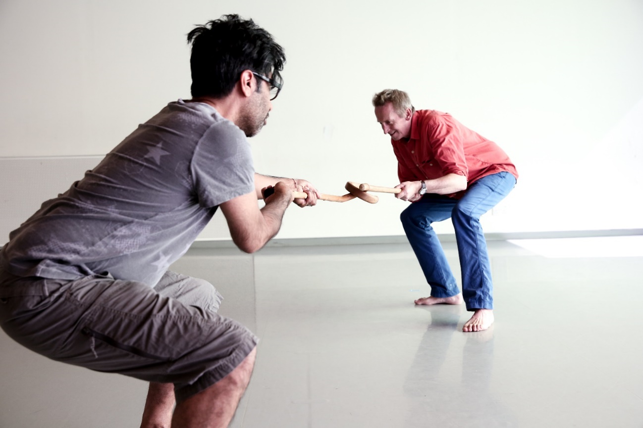 Picture of two men dancing. Each holds the end of piece of wood, puling as if in a tug of war
